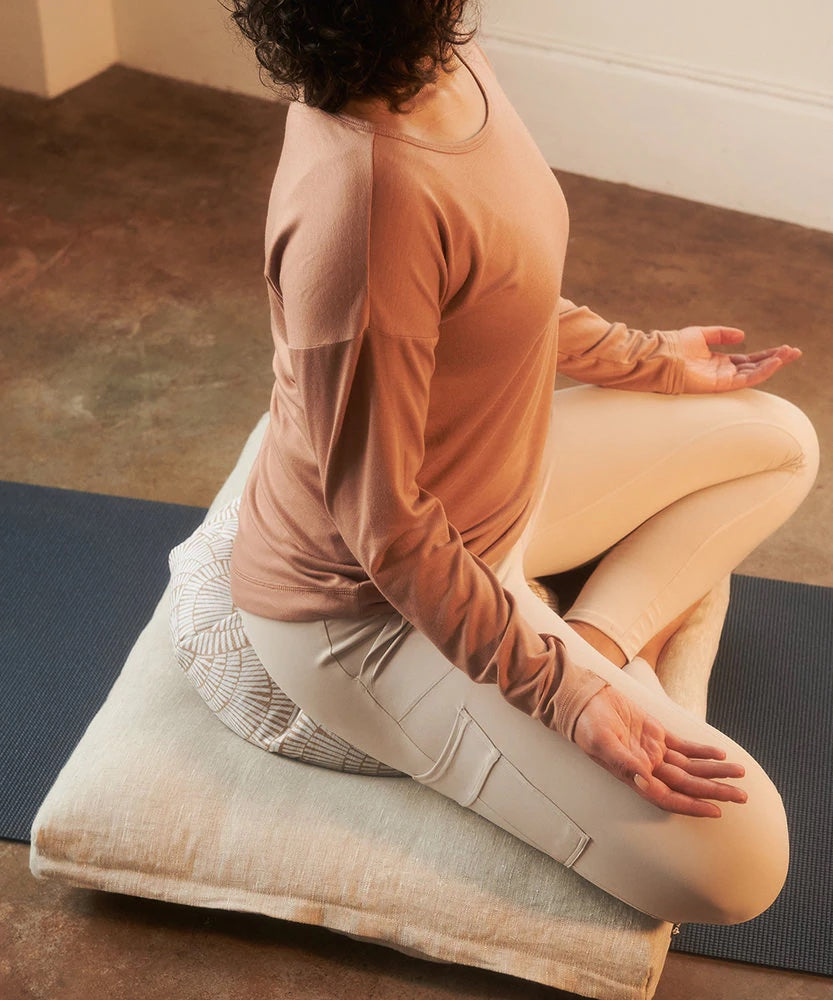 A woman in sukhasana pose sitting on a cushion and yoga mat.