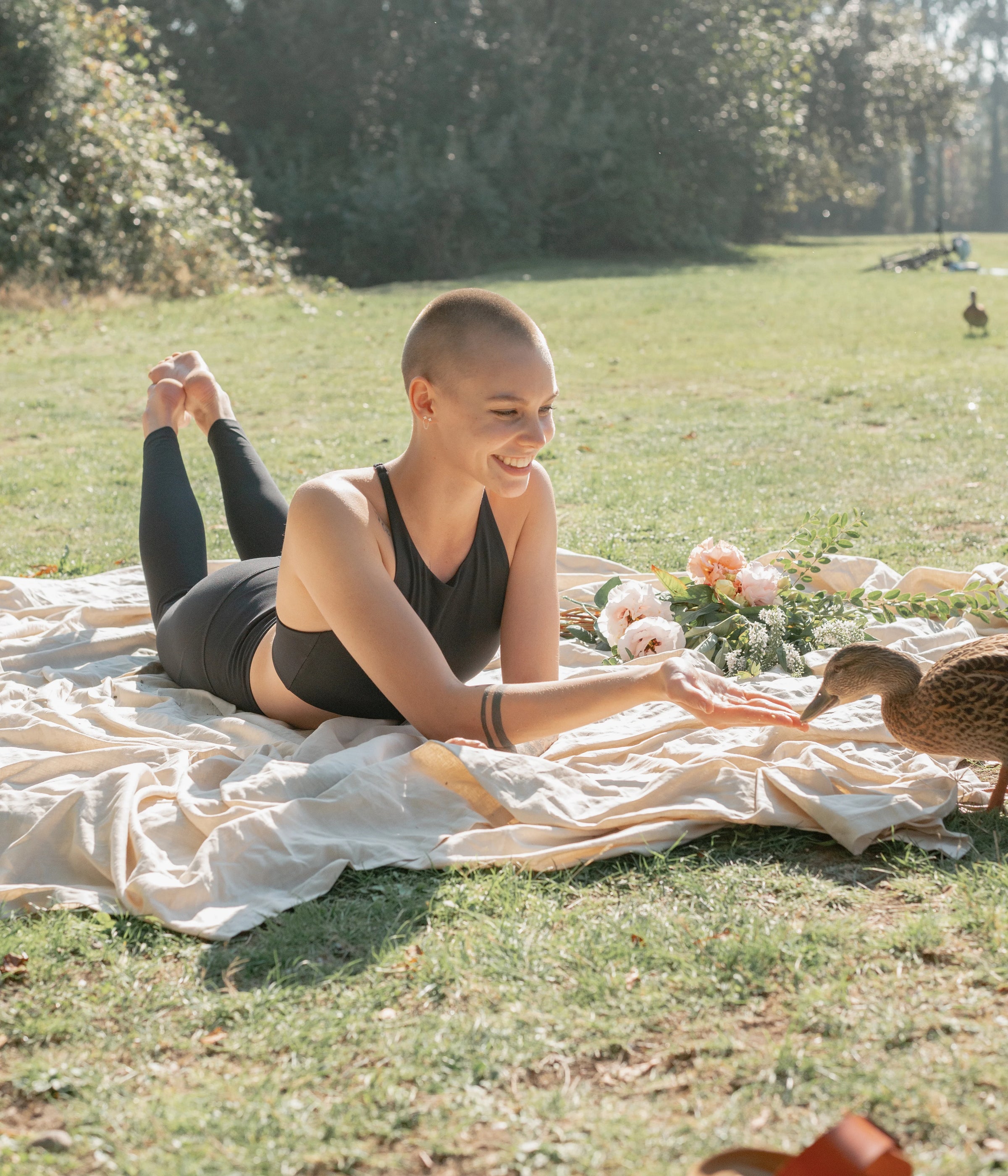Woman in a black sports bra and leggings laying on a blanket outside and feeding a duck.