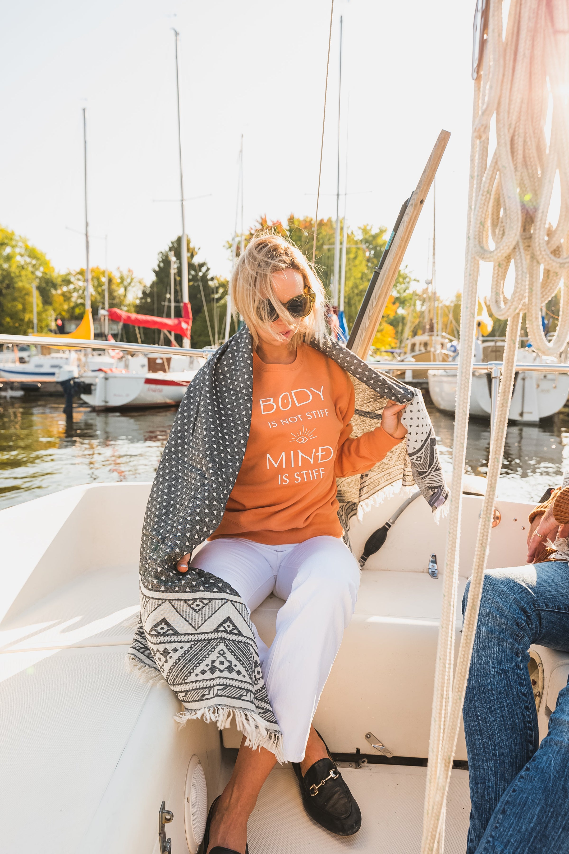 Woman on sail boat with bright orange sweater on and a black and white beach blanket wrapped around her shoulders.