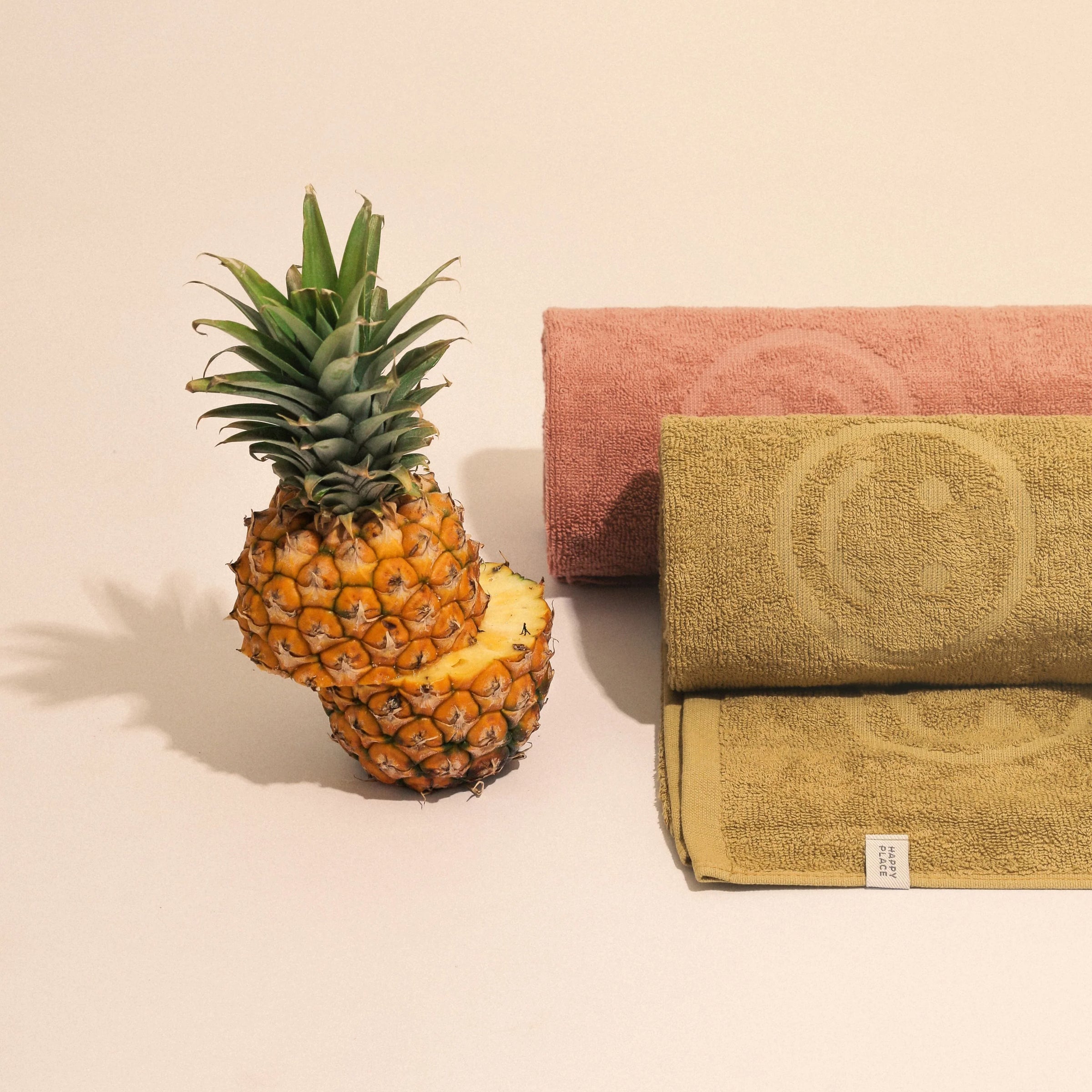 A pink and a green rolled up Happy place towel by a pineapple.
