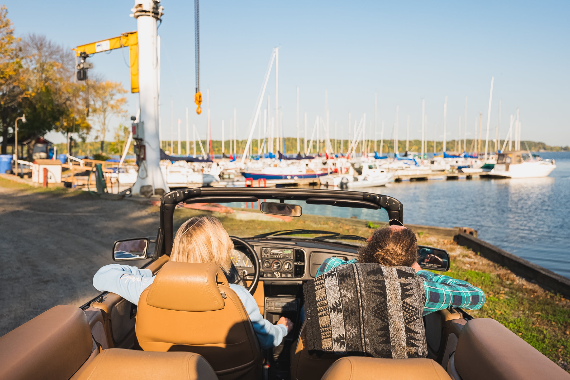 Two people in a convertible facing a dock with a lot of sailing boats.